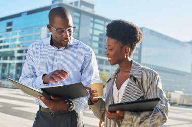 İki profesyonel meslektaş, modern bir şehir ortamında açık havada iş görüşmesi yapıyorlar. Kadın dikkatle dinlerken erkek ayrıntıları açıklayan açık bir klasör tutuyor.