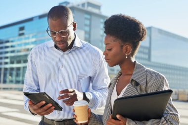 Profesyonel kıyafetli bir erkek ve kadın açık havada bir iş görüşmesi yapıyorlar. Adam ekranı işaret ederken elinde bir tablet tutuyor ve kadın elinde bir kahve fincanı ve dizüstü bilgisayar tutuyor.. 