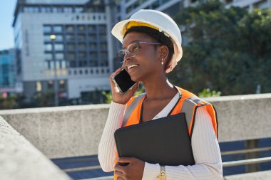 Kasket ve güvenlik yeleği giyen bir kadın inşaat işçisi cep telefonuyla konuşurken şehir ortamında dışarıda duruyor. Elinde bir dosya var ve odaklanmış ve profesyonel görünüyor.