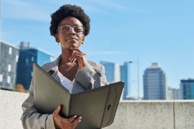 Profesyonel bir kadın, elinde bir klasör, kariyer hırsı ve başarısını simgeleyen bir şehir manzarasında özgüvenle duruyor. Düşünceli ifadesi kararlılığını ve dikkatini gösteriyor.