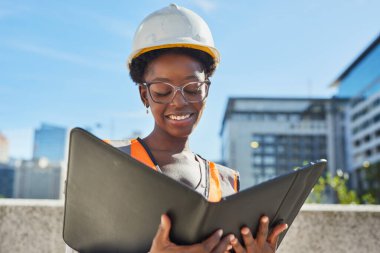 Kendine güvenen kadın mühendis inşaat alanındaki belgeleri incelerken çelik yelek ve şapka takıyor. Arka plandaki şehir manzarası kentsel ortamı vurgular.
