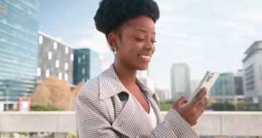 Şık bir paltosu olan genç siyahi bir kadın şehrin iş bölgesinde duruyor ve akıllı telefonunu kullanırken gülümsüyor. Kentsel çevrede bir anlık bağlantı ve verimliliğin tadını çıkarıyor..
