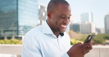 Mutlu bir Afro-Amerikan işadamı şehirdeki modern cam ofis binalarının önünde telefonuna gülümsüyor. Mavi ve beyaz çizgili bir gömlek giyiyor ve akıllı telefon kullanıyor.. 