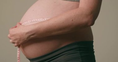 Hamile bir kadın göbeğini mezurayla ölçüyor. Karnındaki şişliği ölçen bir kadına yakın çekim. Hamilelik, annelik, beklenti, aşk, aile kavramı..