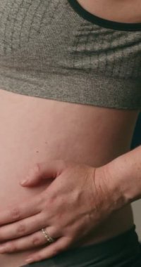 Hamile bir kadın karnını ve yoga minderini tutar. Gri spor sutyen ve gri tayt giyiyor. Sağlıklı hamilelik ve zindelik kavramı. Yavaş çekim..