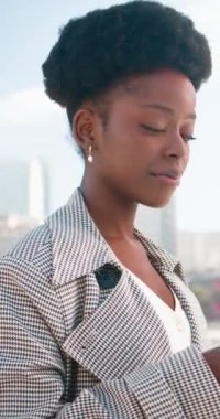 İş kıyafetleri içinde genç bir siyahi kadın akıllı telefonunu çatıda dikilirken kullanıyor. Şehir manzarasına bakıyor. Modern binalar başarılı bir iş kariyeri öneriyor.