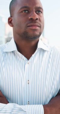 Genç, siyah bir işadamı kollarını kavuşturmuş, düşünceli ve kararlı bir şekilde şehirde duruyor. Kurumsal bir ortamda hırs, başarı ve liderliği somutlaştırıyor..