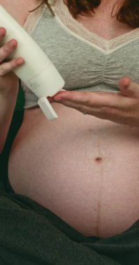 Hamile bir kadın yatağına oturur ve karnına losyon sürer. Gri bir sutyen ve gri pijama pantolon giyiyor. Arka planda yeşil bir duvar ve bir saat ve bitki var..