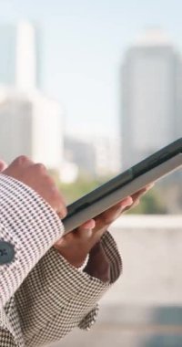 Bir iş kadınının elleri, şehir siluetinin arka planına karşı tablet kullanırken yakın plan görüntüsü. Bu sahne iş, teknoloji ve şehir yaşamını çağrıştırıyor.