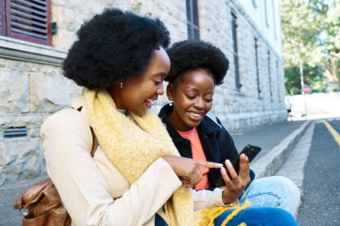 İki gülen arkadaş şehrin kaldırımında oturmuş akıllı telefona bakarken eğleniyor. Sıcak, güneşli bir gün onların neşeli ruh hallerine neşe katıyor, arkadaşlık ve mutluluk temalarını vurguluyor..