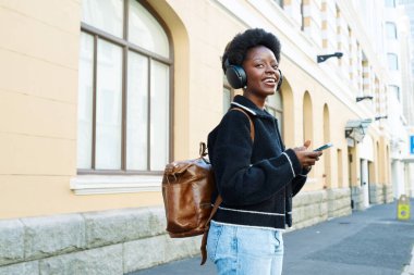 Kulaklık takan ve elinde akıllı telefon tutan neşeli genç bir kadın şehir caddesinde parlak bir gülümsemeyle yürüyor. Sırt çantasıyla gününün ve müziğinin tadını çıkarıyor..