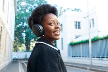 Neşeli bir yüzü olan genç bir kadın şehir ortamında yürürken kulaklıkla müzik dinliyor. Parlak gülüşü ve şık kıyafeti mutluluğu ve özgüveni yansıtıyor.. 