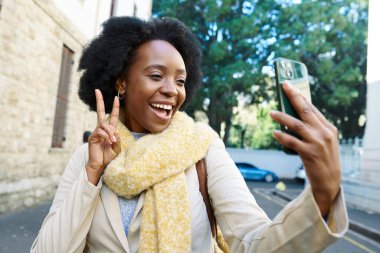 Sarı eşarplı neşeli bir kadın açık havada selfie çekerken ışıl ışıl gülümsüyor. Canlı bir şehir ortamında bir anlık mutluluk yakalayan bir barış işareti gösteriyor.