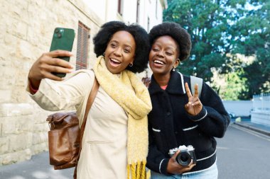 İki neşeli arkadaş güneşli bir şehir macerasında neşeli bir selfie çekiyorlar. Bir arkadaş cep telefonu tutarken, diğeri gençlik enerjisini somutlaştıran bir barış işareti gösteriyor.