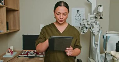 Kliniğinde tablet kullanan bir optometrist. Göz doktoru hasta dosyalarını inceler, randevularını ayarlar ve kliniğini dijital tabletinden yönetir. Optometri, sağlık, teknoloji.