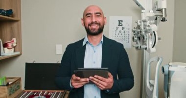 Gülümseyen Optometrist Göz Kliniği 'nde Dijital Tablet kullanıyor. Arka planda göz çizelgesi ve ekipmanı olan dost canlısı bir doktor. Profesyonel Sağlık ve Vizyon Bakımı Konsepti.