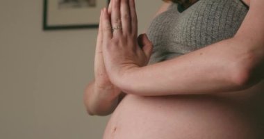 Elleri dua pozisyonunda meditasyon yapan hamile bir kadının karnına yakın çekim. Gri spor sutyen ve gri yoga pantolonu giyiyor. Konsept gebeliği, farkındalığı ve barışı aktarır.. 