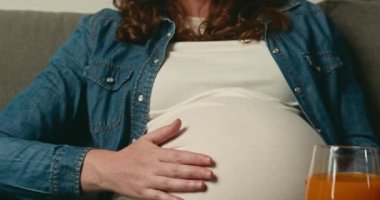 Hamile bir kadın kanepeye oturur ve karnını nazikçe ovar. O bir kot gömlek ve beyaz bir tişört giyiyor. Yanında bir bardak portakal suyu var..