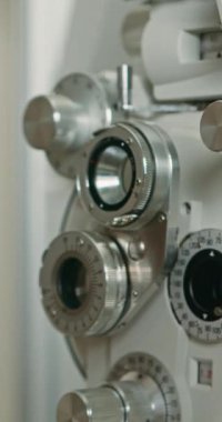 Göz muayenesi için kullanılan, göstergeler ve lenslerle bir optometri makinesinin yakın çekimi. Odak bir göz grafiğine kayar, görme testinin araçlarını ve sürecini vurgular.