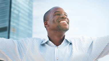 Profesyonel olarak giyinmiş neşeli bir birey açık havada duruyor, arka planda modern binalarla berrak bir gökyüzünün altında ışıldıyor, pozitiflik, güven ve kentsel başarı hissi veriyor..