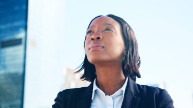 Kendine güvenen Afrikalı Amerikalı iş kadını resmi giyinmiş yukarı bakıyor, modern şehir manzarasına karşı duruyor..