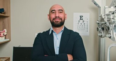 Ofisinde duran kendinden emin bir optometristin portresi. Kollarını kavuşturup gülümseyerek profesyonellik ve göz bakımında uzmanlık yayıyor. Arkaplanda bir göz çizelgesi var.
