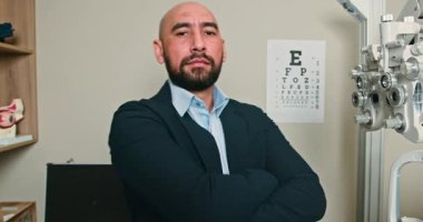 Kendine güvenen bir optometrist kollarını kavuşturup doğrudan kameraya bakıyor. Arka planda, bir göz çizelgesi ve göz teçhizatı görünür. 