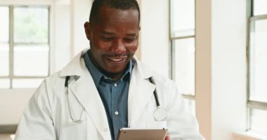 Dost canlısı bir erkek doktor, tabletindeki hasta bilgilerini incelerken gülümsüyor. O bir hastane koridorunda duruyor. Doktor beyaz önlük ve steteskop giyiyor ve mutlu görünüyor..