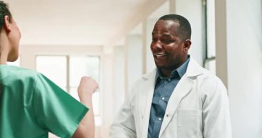Bir erkek doktor ve bir kadın hemşire hastane koridorunda yumruk tokuşturuyor. İkisi de gülümsüyor ve birbirlerini görmekten mutlu görünüyorlar. Doktor hemşireyle konuşuyor..