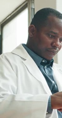 Ciddi bir Afrikalı Amerikalı tıp doktoru hasta bilgilerini tabletinden inceliyor. Kaydırırken endişeli görünüyor. Koyu mavi bir gömleğin üzerine beyaz bir laboratuvar önlüğü giyiyor..