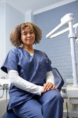 Gülümseyen diş hekimi bir dişçi kliniğinde oturuyor. Görüntü profesyonelliği ve iyi ışıklandırılmış ve modern bir klinik ortamının yer aldığı oral sağlık hizmetlerine odaklanmayı gösteriyor..