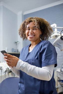 Mavi önlük giymiş neşeli bir diş hekimi modern bir diş kliniğinde dururken akıllı telefon kullanıyor. Sahne profesyonellik, teknoloji ve olumlu bir çalışma ortamı sunuyor..