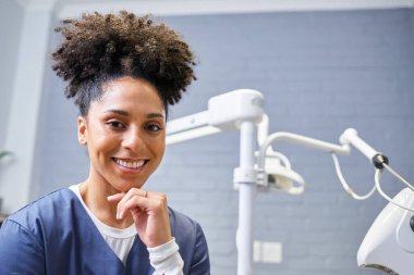 Neşeli bir diş hekimi, çağdaş bir diş muayenehanesinde güven ve beceri yayarak resmedilir. Diş araçları arka planda görünür, klinik ayarları vurguluyor.