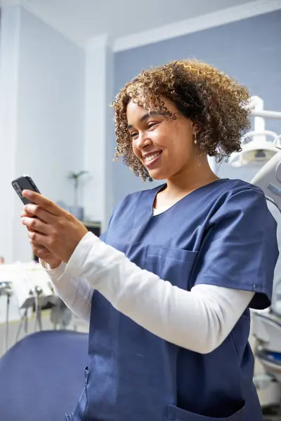 Mavi önlük giyen bir sağlık çalışanı akıllı telefonunu tutarken gülümsüyor. Arka plan modern bir diş muayenehanesini gösteriyor, profesyonelliği ve teknolojiyi vurguluyor..