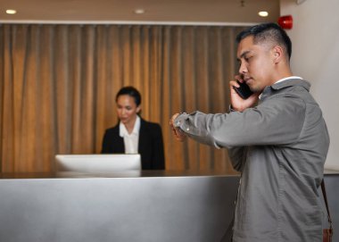 İş kıyafeti giymiş bir gezgin otel kayıt gişesinde telefonla konuşurken saatine bakmış. Arka planda, bir resepsiyonist dikkatle ayakta duruyor..