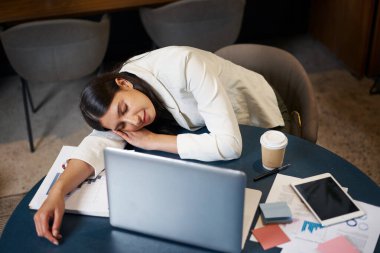 Yorgun, profesyonel bir kadın kafasını dizüstü bilgisayar, çizelge ve kahveyle kaplı bir masanın üzerinde dinlendiriyor. İşyeri yorgunluğunun, aşırı çalışmanın ve dinlenme ihtiyacının özünü yakalar.