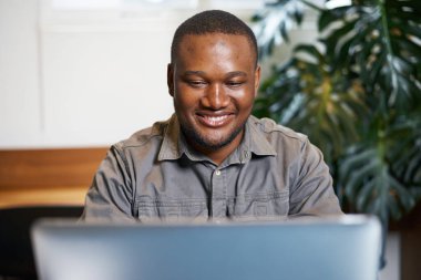 Bir adam parlak bir ofis ortamında dizüstü bilgisayarıyla çalışırken gülümsüyor. Atmosfer profesyonel ama rahat, yeşil bitkiler üretkenliğe olanak sağlayan hoş ve taze bir çevre yaratıyor..