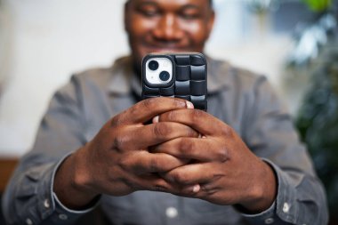 Bir kişi şık bir çantayla akıllı bir telefon tutuyor, modern bir ortamda nişanlı ve mutlu görünüyor. Resim bağlantı, iletişim ve keyif temalarını iletir.