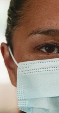 Yüz maskesi takan genç bir kadının yakın çekimi. Kahverengi gözleri doğrudan kameraya bakıyor. Burnunun ve alnının köprüsünde hafif çiller var. Yüzü ciddi ve düşünceli..