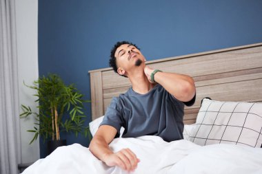 Adam yatakta boynunu tutarak rahatsızlığını dile getiriyor. Yatak odasında modern bir dekor tarzı var. Mavi aksanlı bir duvar ve ahşap bir yatak çerçevesi var. Sakin ve rahat bir atmosferi çağrıştırıyor..