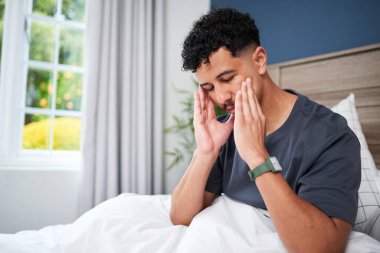 Bir adam iyi ışıklandırılmış bir yatak odasında oturur, başını görünür bir stres ya da rahatsızlık içinde, pencerenin yanında ve beyaz perdelerin yanında. Görüntü, hayal kırıklığı, yorgunluk ya da tefekkür gibi duyguları iletir..