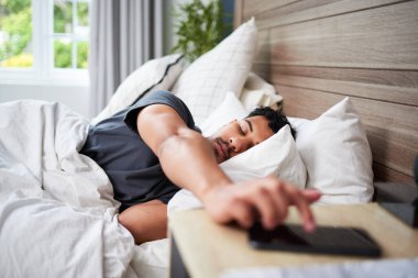 Yatakta rahatça dinlenen genç bir adam çalar saati ayarlamak için elini uzatır. Yatak odasında sakin bir sabah ortamı var. Doğal ışık ve lüks yatak örtüsüyle vurgulanıyor..