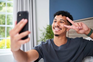 Kıvırcık saçlı neşeli genç bir adam akıllı telefonuyla selfie çekerken barış işareti yapıyor. Sahne rahat ve aydınlık modern bir ev ortamında geçiyor..