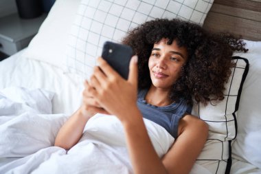 Kıvırcık saçlı genç bir kadın yatakta rahat, akıllı telefonuyla meşgul. Görüntü, rahat bir ev ortamında sessiz bir rahatlık ve dijital bağlantı hissi yayıyor..