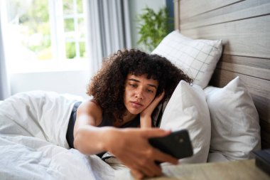 Kıvırcık saçlı genç bir kadın akıllı telefonuna bakarken, parlak ve modern bir yatak odasında bir yatakta uzanıyor. Sahne rahatlama, kişisel bağlantı ve evdeki huzurlu yaşam tarzını aktarır..