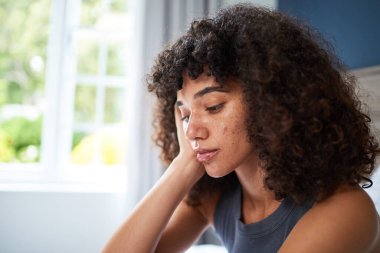 Pencerenin yanında tefekküre dalmış genç bir kadının yakından görüntüsü. İfadesi düşünceliliğini yansıtıyor ve doğal bukleleri odaya giren yumuşak, çevresel gün ışığıyla aydınlanıyor..