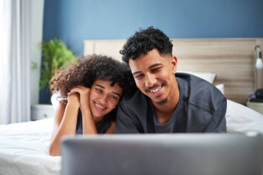 Genç ve mutlu bir çift, bir cihazın içeriğini izleyerek rahatça yataklarında uzanıyorlar. Gülüşleri ve gülüşleri, bu samimi aile ortamında paylaştıkları sıcaklığı ve bağlantıyı gösteriyor..