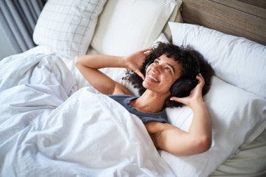 Neşeli bir kadın, rahat beyaz çarşafların altında uzanmış siyah kulaklıklarla müzik dinliyor. Rahatlamış duruşu ve ışıl ışıl gülüşü, bu huzurlu kapalı mekan keyfi anındaki memnuniyetini yansıtıyor..