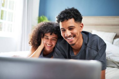 Neşeli bir çift, dizüstü bilgisayarlarında içeriklerini incelerken gülümseyerek yataklarında kaliteli zaman geçiriyorlar. Sahne, mutluluk, bağ ve konforu parlak ve huzurlu bir ev ortamında yakalıyor..