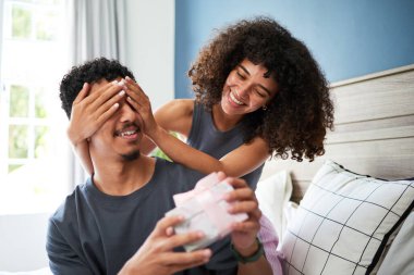 Sıcak evlerinde güneşli bir günde içten bir sürpriz hediyeyle eğlenen genç bir çiftin neşeli ve samimi anı. Atmosfer neşe, kahkaha ve sevgi dolu..